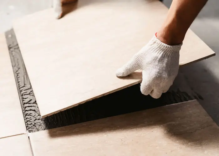 Hand mit weißem Handschuh legt eine Fliese auf verlegten Fliesenkleber auf Bodenfliesen.