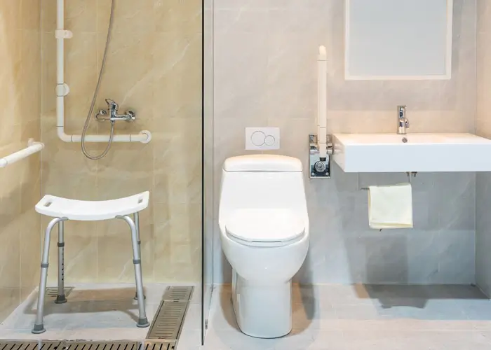 Barrierefreies Badezimmer mit Duschstuhl, Haltegriffen, Toilette und Waschbecken mit Handtuchhalter.