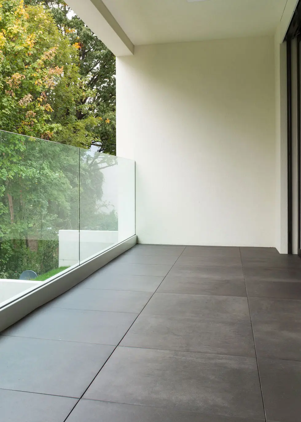 Moderner Balkon mit großformatigen grauen Bodenfliesen und Glasgeländer, im Hintergrund Bäume sichtbar.