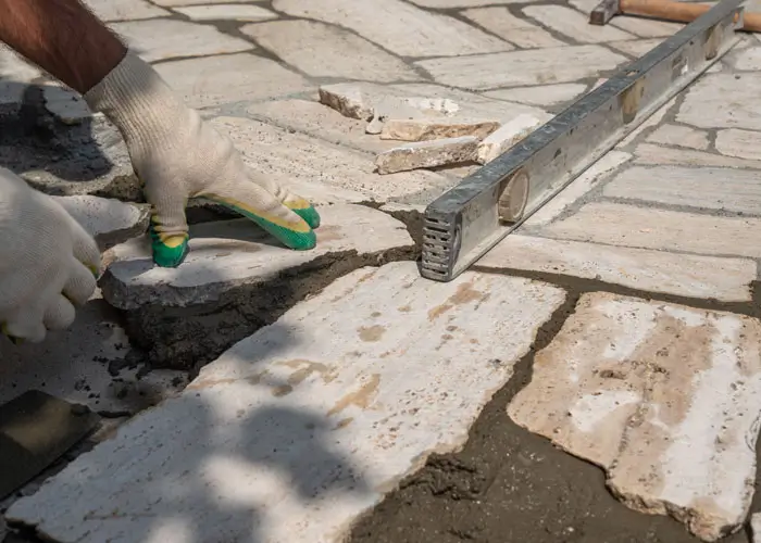Hände mit Arbeitshandschuhen verlegen Natursteinplatten auf frischem Mörtel neben einer Wasserwaage.