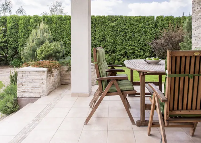 Terrasse mit Holztisch und Stühlen mit grünen Polstern, umgeben von Hecke und Steinmauer.