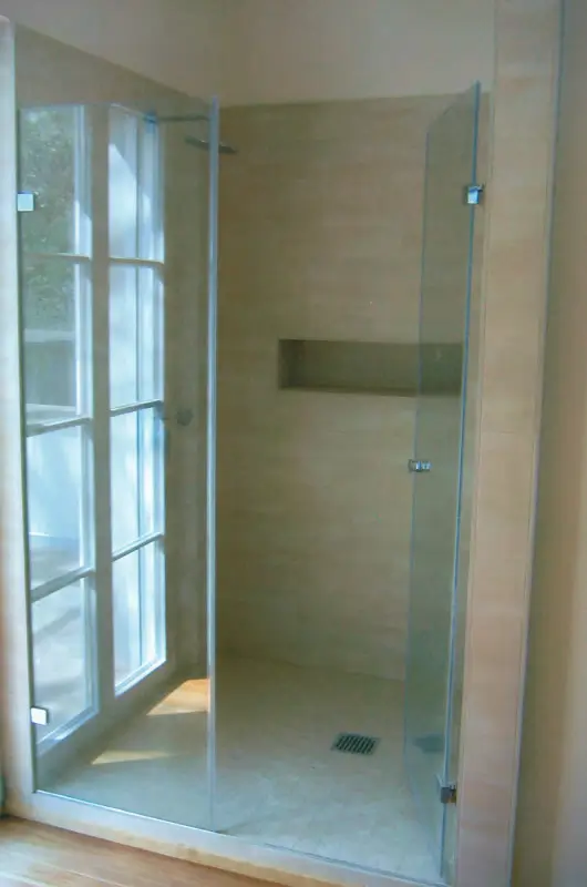 Dusche mit rahmenloser Glasabtrennung und eingebautem Wandfach neben einem Fenster mit weißen Rahmen.